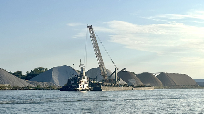 A wide-angle photo showing a tug next to an empty barge near land. On shore are three large hills of rock, plus three large dome-shaped silos. A crane on the empty barge is pointed toward shore in preparation for loading.
